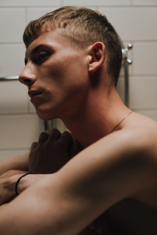 Free Young man with short hair sitting thoughtfully in a modern tiled bathroom. Stock Photo