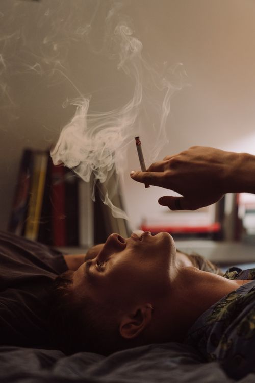 Free A man lies indoors, blowing smoke, creating a relaxed, moody atmosphere. Stock Photo