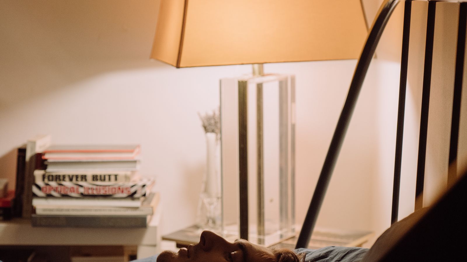 A man resting on a bed with his eyes closed in a warmly lit bedroom during the evening