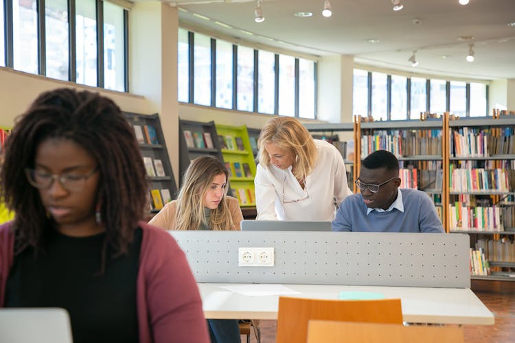 Young Diverse Students Coworking On Laptop In Library With Assistance Of Female Teacher