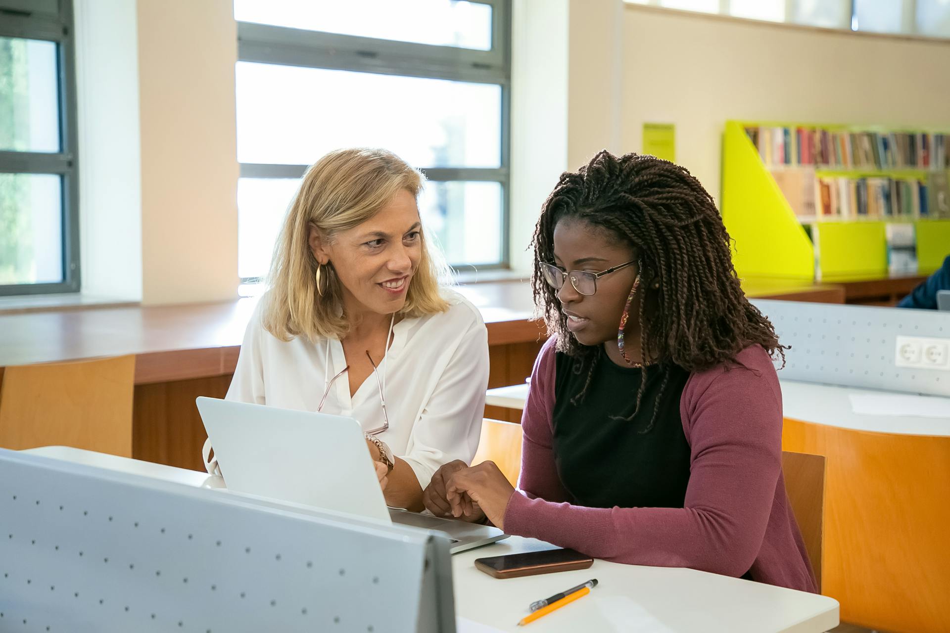 Positive adult female teacher explaining task to young black student with Afro braids doing assignment on laptop in modern classroom