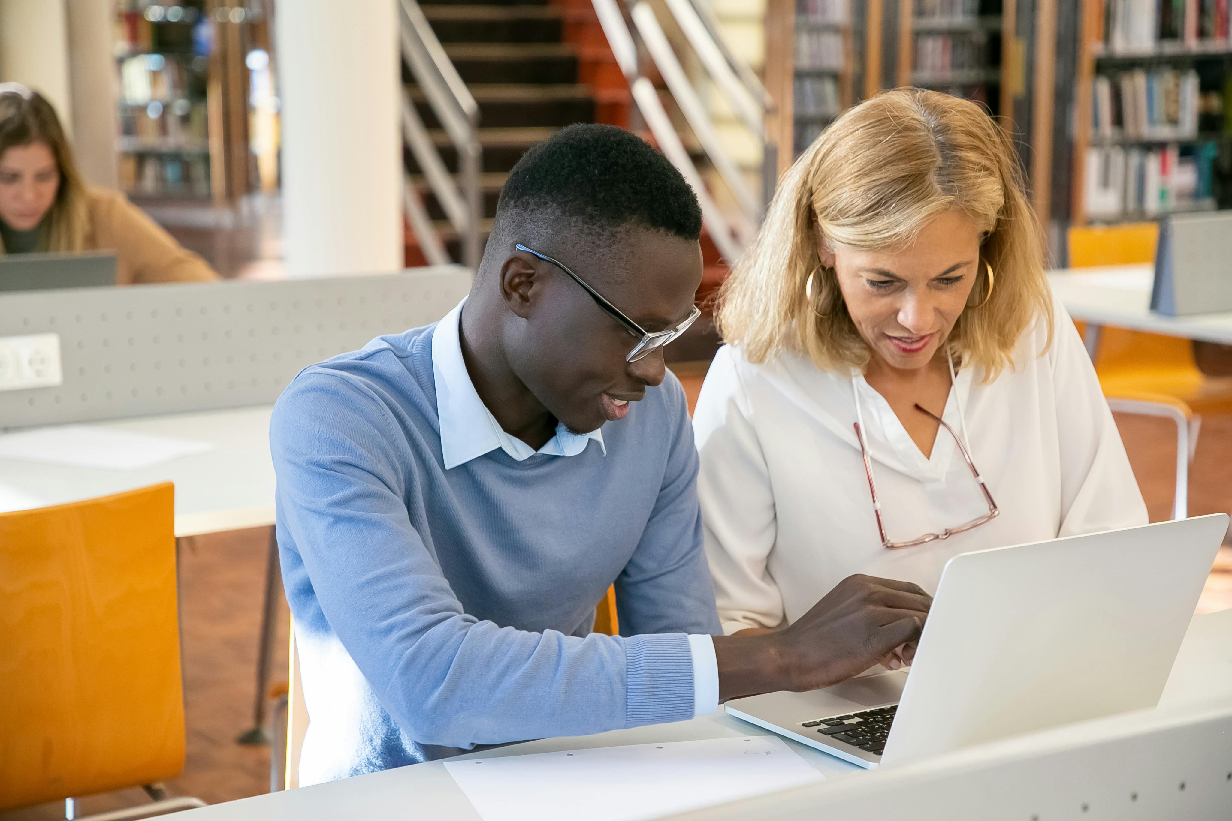 female teacher helping ethnic male student working on laptop in library