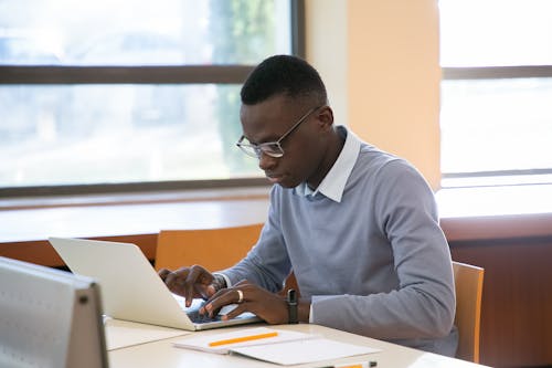 Concentrated young African American male student in formal clothes and eyeglasses reading notes in planner and typing on laptop while doing assignment in modern workspace