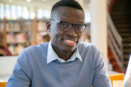 Free Positive African American male learner with toothy smile in eyeglasses sitting in library Stock Photo