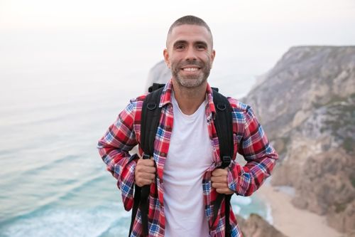 Free A smiling man in a plaid shirt hiking with a backpack on Portugal's scenic cliffs. Stock Photo
