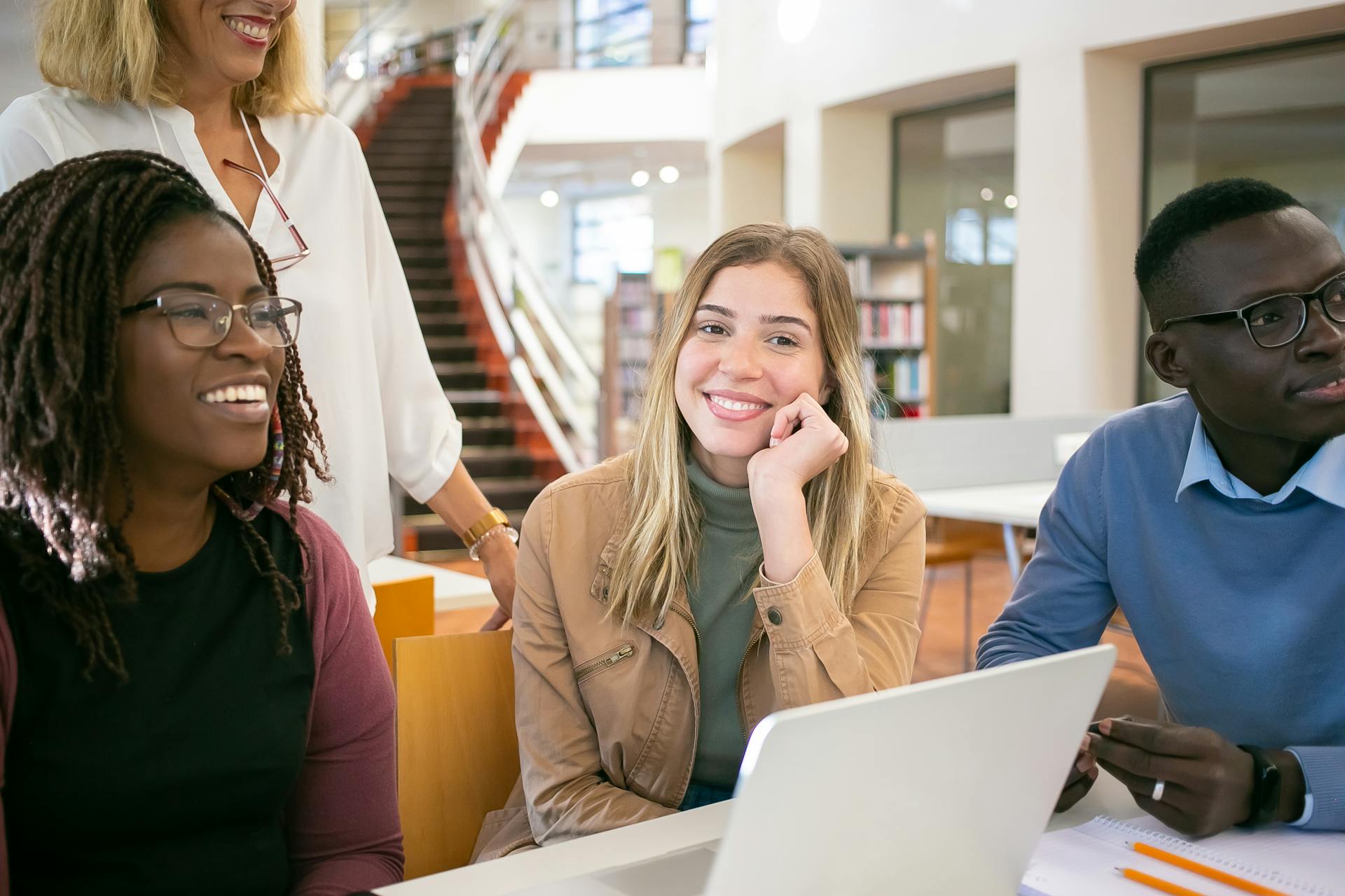 Cheerful multiracial young coworkers sitting at table with netbook and discussing project with teacher in university