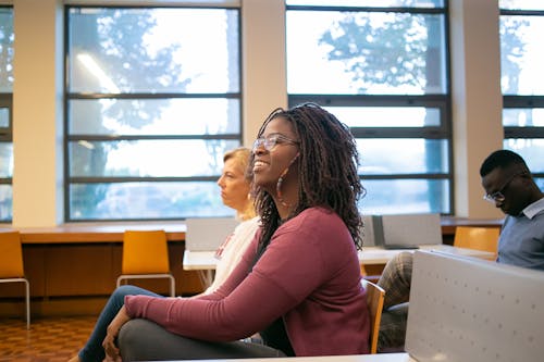 Side view of positive African American female student sitting on chair near professor and classmate while listening information attentively