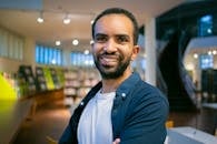 Smiling ethnic bearded student with toothy smile standing against bookshelves in library in college