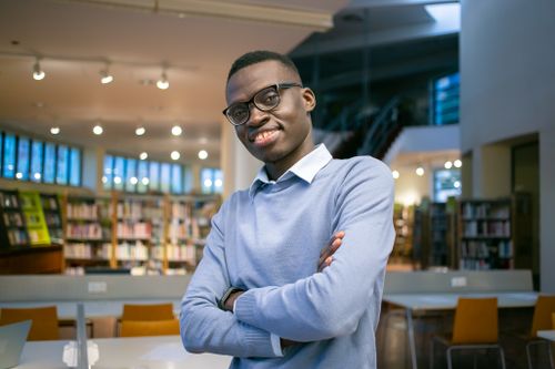 Free Positive African American student with crossed arms standing among tables and chairs in library Stock Photo