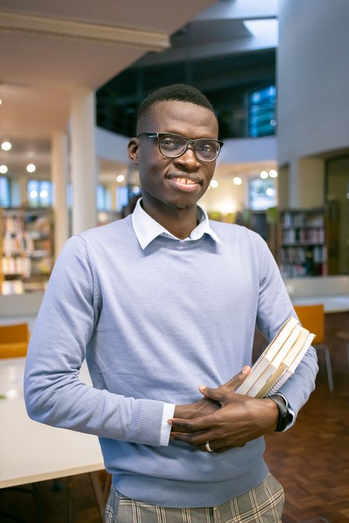 Free Optimistic African American man in eyeglasses with stack of books in hands standing in university library Stock Photo