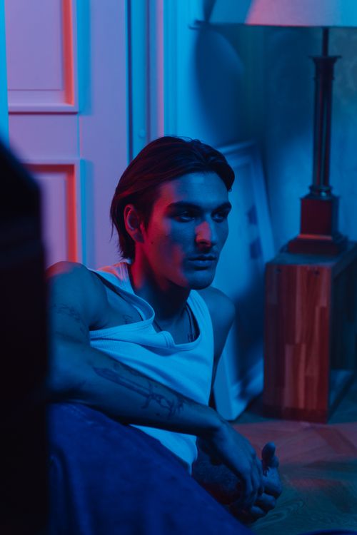 Free A young tattooed man in a tank top sits indoors under creative blue and red lighting effects. Stock Photo