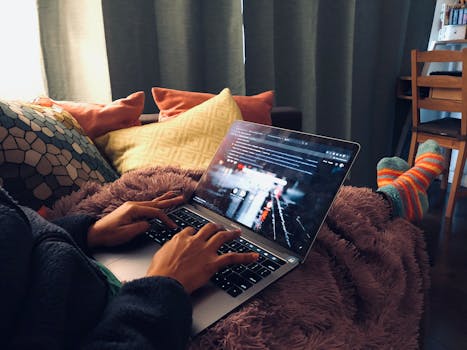 A person relaxing at home using a laptop on a cozy couch with soft lighting.
