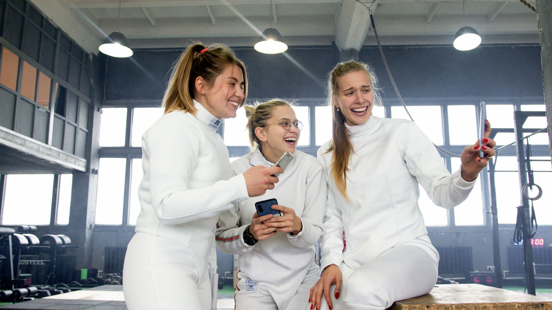 Three women fencers in white uniforms laughing and taking a selfie indoors.