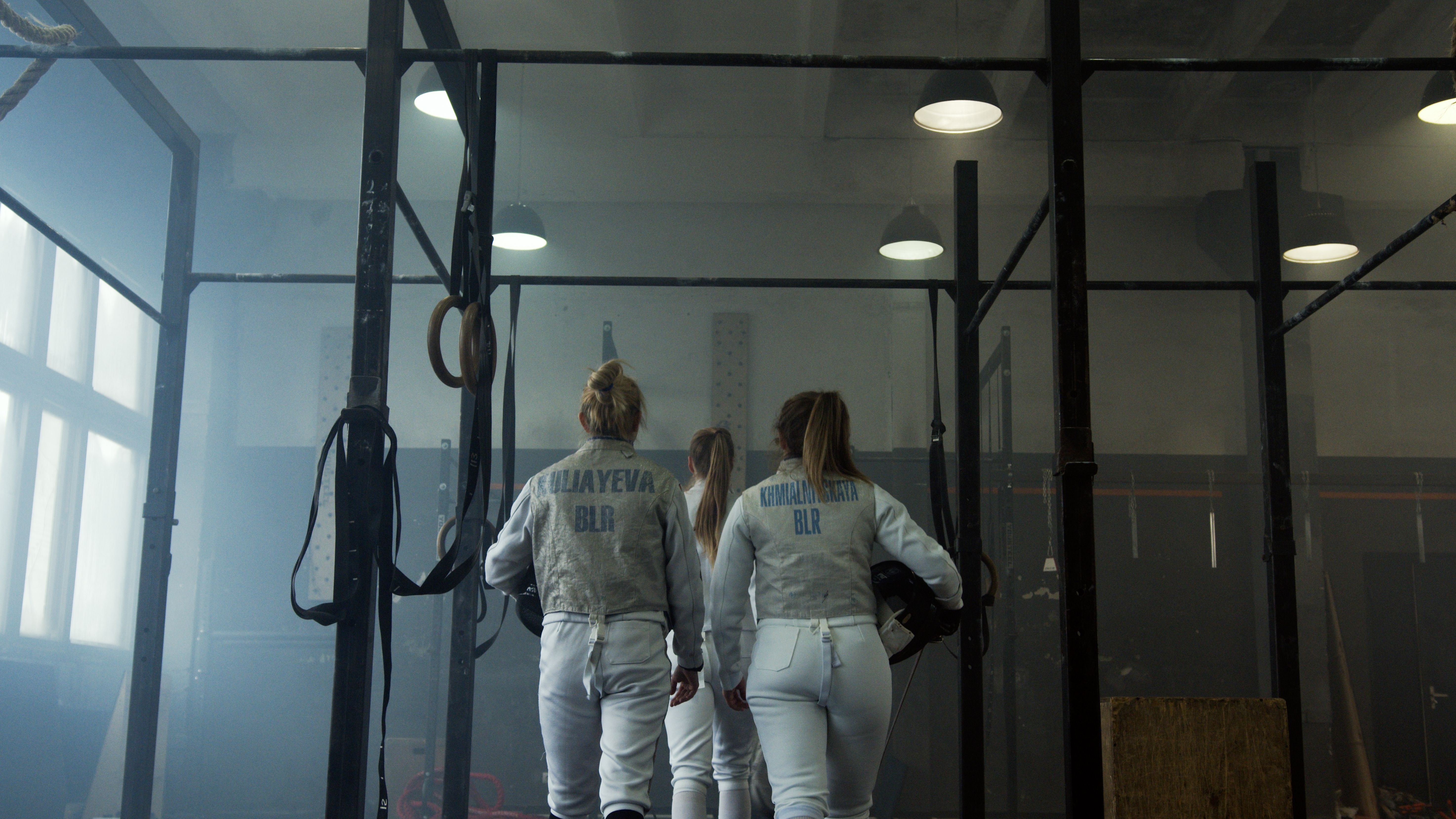 Three female fencers in uniform enter the indoor gym, ready for an intense practice session.