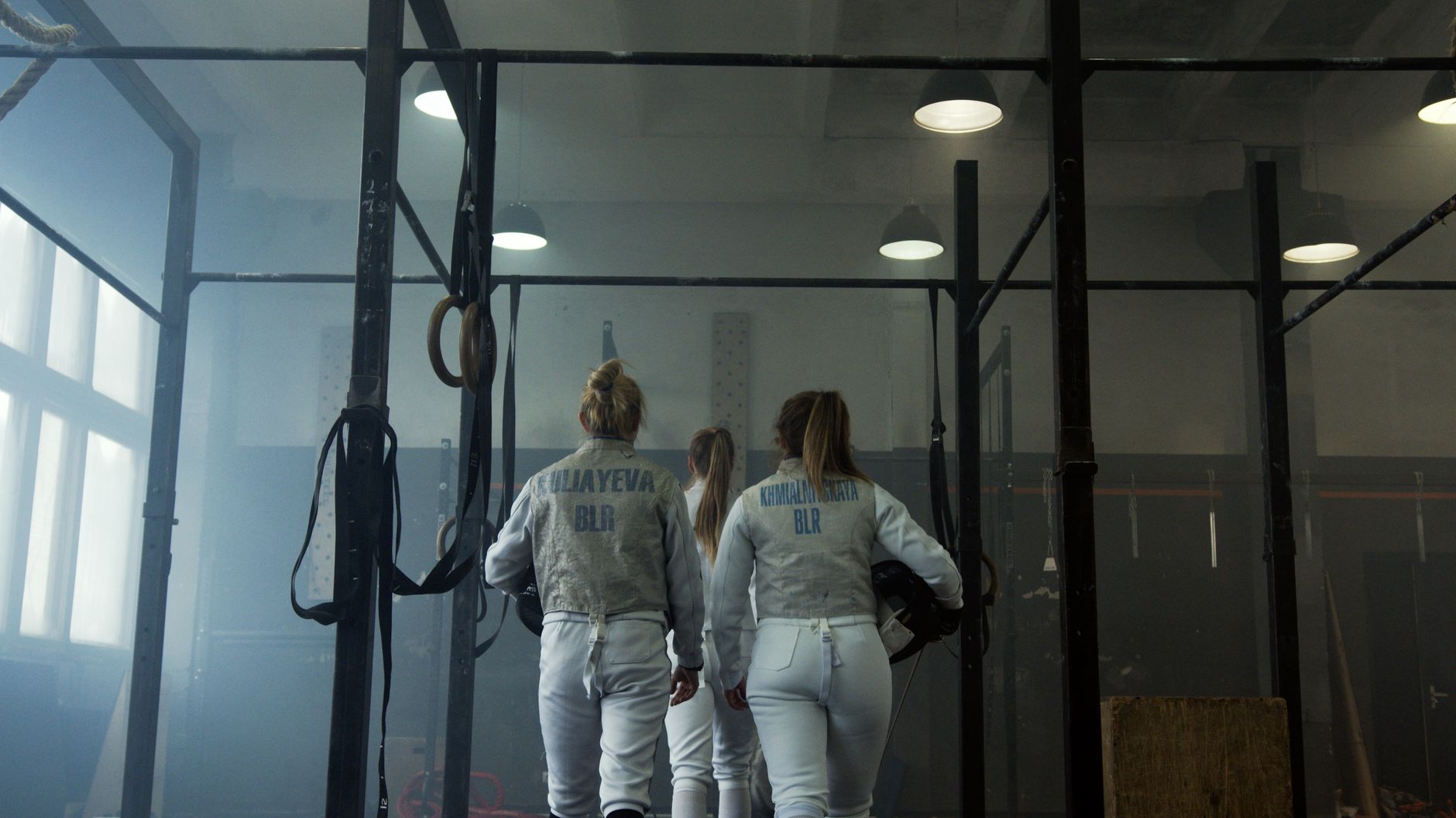 Three female fencers in uniform enter the indoor gym, ready for an intense practice session.