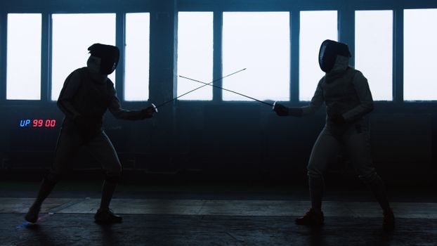 Two athletes engaged in a dynamic fencing duel inside a large hall.