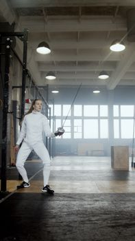 Female fencer in protective gear practicing in a spacious indoor gym setting.