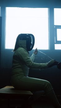 Side view of a female fencer in gear sitting indoors with an epee sword.