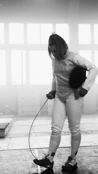 Black and white image of a female fencer holding a fencing mask indoors.
