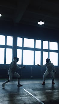 Two women engaged in a dynamic fencing duel indoors, capturing intense competition.