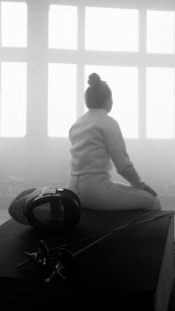 Young female fencer sitting with equipment in a misty room, reflecting mentally after training.