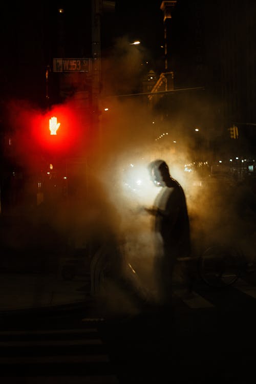 Full length of unrecognizable pedestrians silhouettes walking in white steam on asphalt road on crosswalk near sidewalk and buildings in city district at night