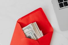 Top view of dollar bills in a red envelope next to a laptop on a white desk.