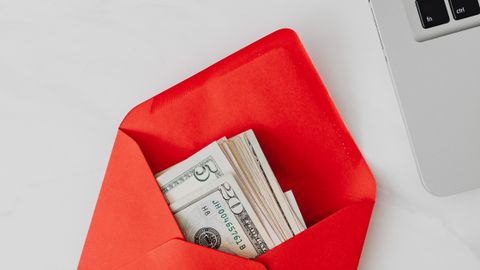 Top view of dollar bills in a red envelope next to a laptop on a white desk.