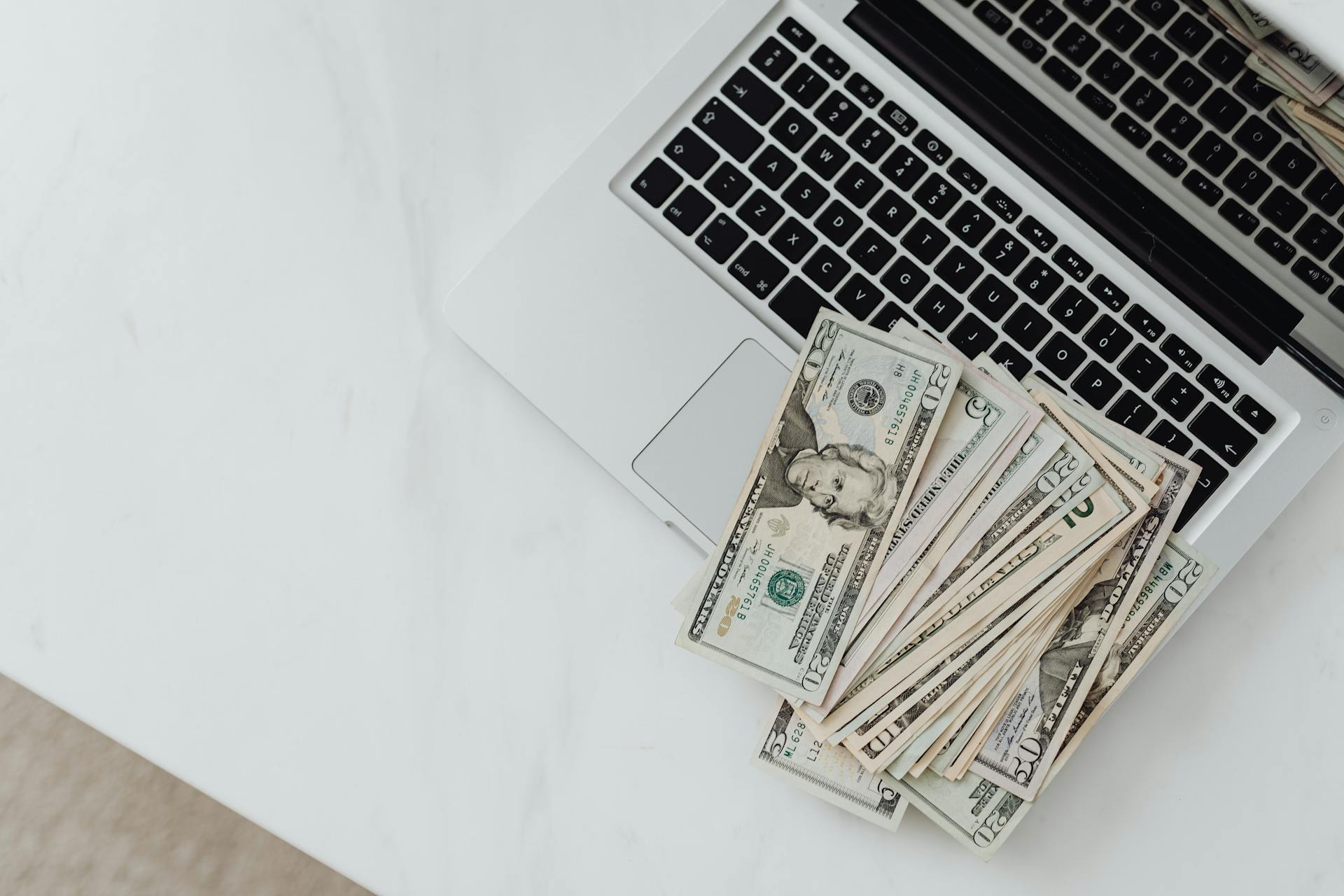 Close-up of US dollar bills scattered on a laptop keyboard on a white desk, symbolizing digital finance.