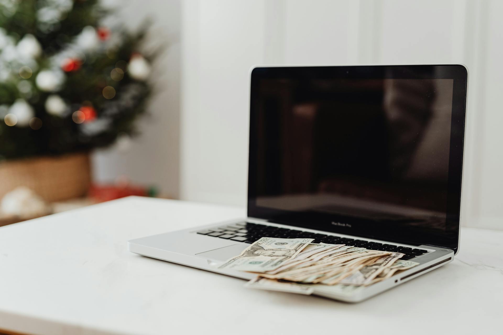 A laptop on a desk covered with cash, featuring a Christmas tree in the background.