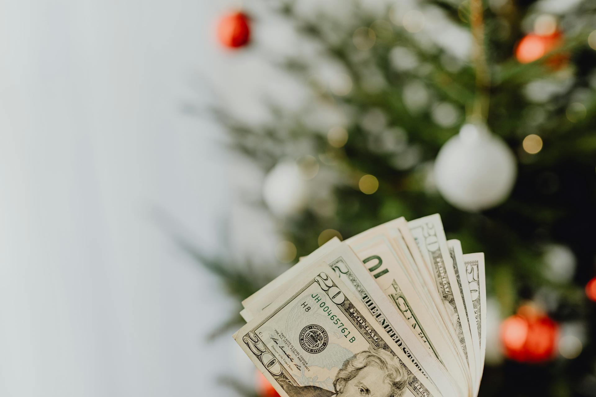 Stack of US dollar bills in front of a decorated Christmas tree, symbolizing holiday spending.