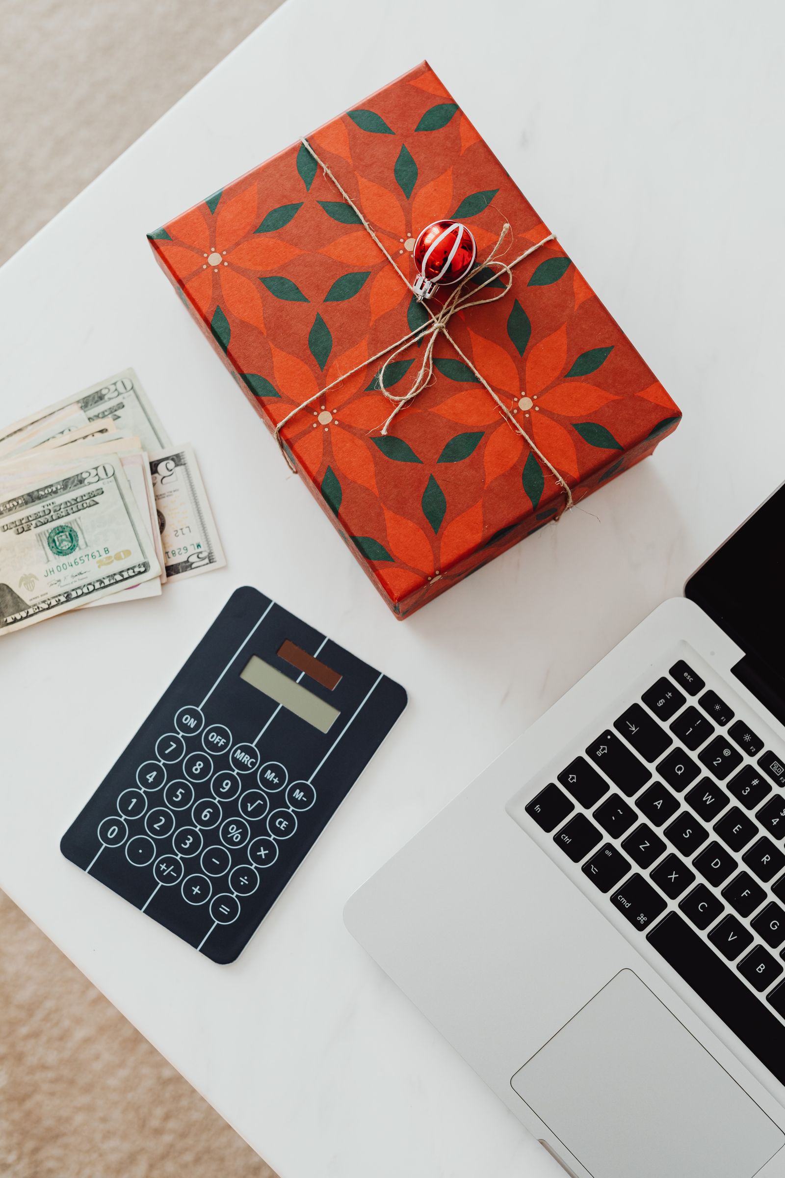Wrapped Christmas present beside cash, a calculator, and a laptop on a white table.