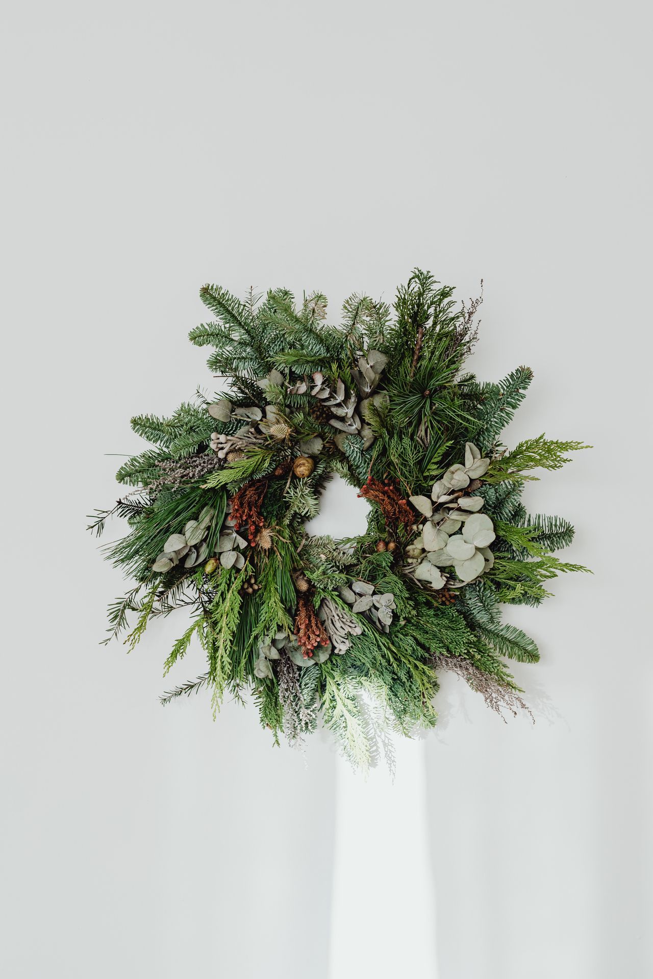 Natural handmade wreath of mixed evergreens, eucalyptus, dried grasses, and pinecones on a white wall.