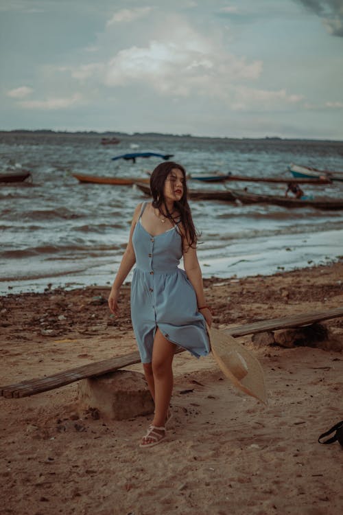 Free Full body of charming female with long dark hair in summer dress standing on sandy coast near wavy ocean in windy day Stock Photo