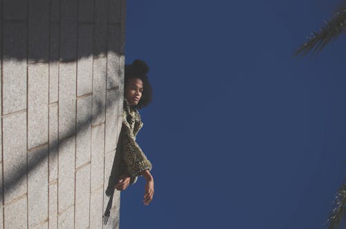From below of young African American female in warm clothes looking at camera while leaning on stone fence under blue cloudless sky in sunny day