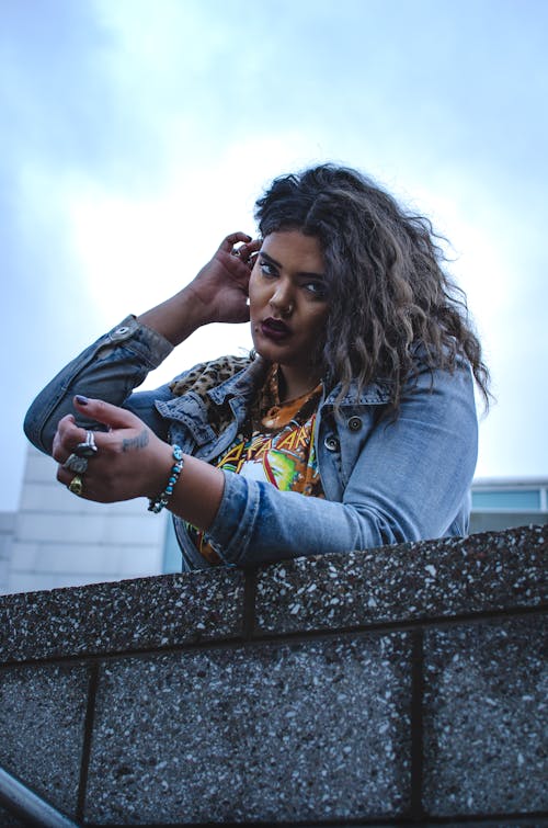 From below of young serious ethnic female in casual clothes leaning on stone barrier in city street under blue sky and looking at camera