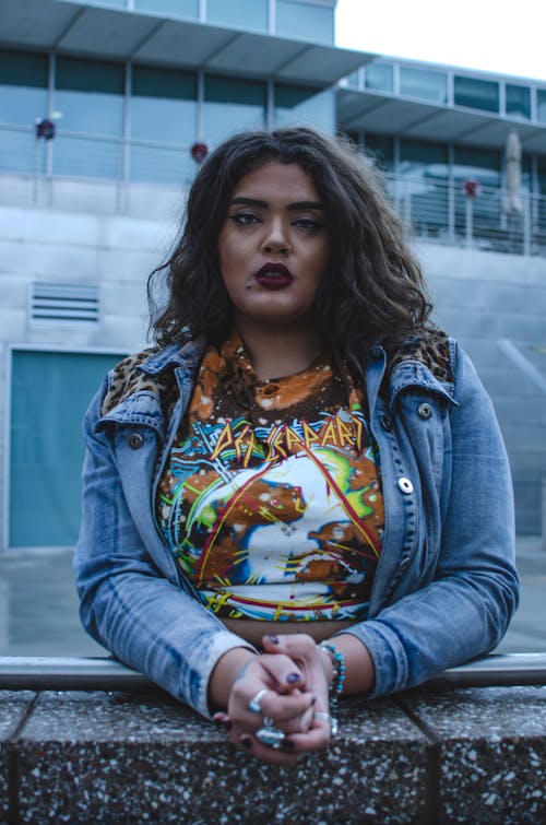 Young serious ethnic plus size woman in casual outfit standing near stone barrier in city street and looking at camera