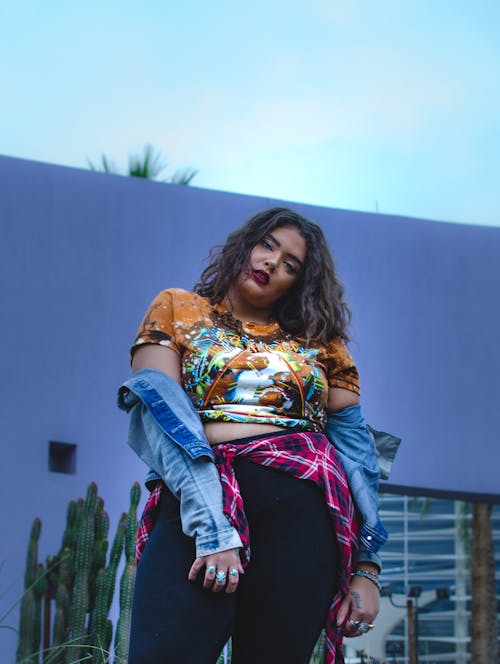 Young confident ethnic plus size female in casual clothes standing near blue wall in city street and looking at camera under blue cloudless sky