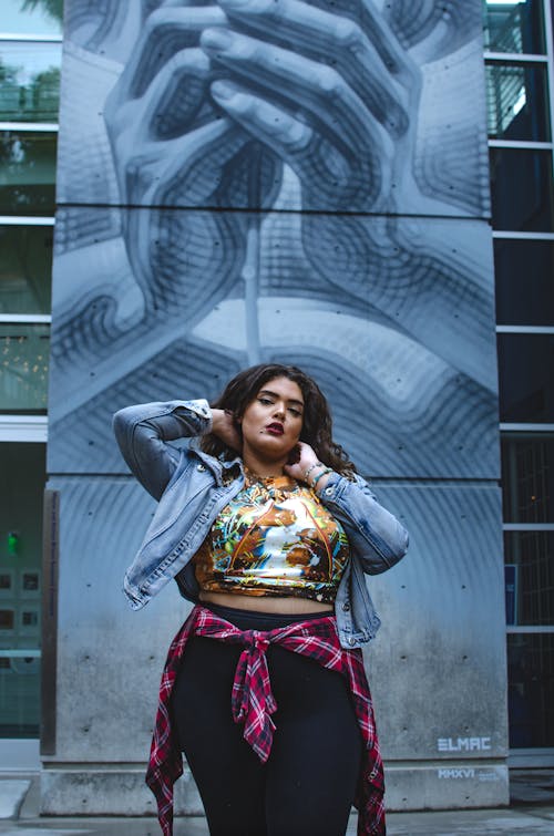 Young confident ethnic body positive female in casual clothes standing near wall with blue graffiti in city street and looking at camera in daytime