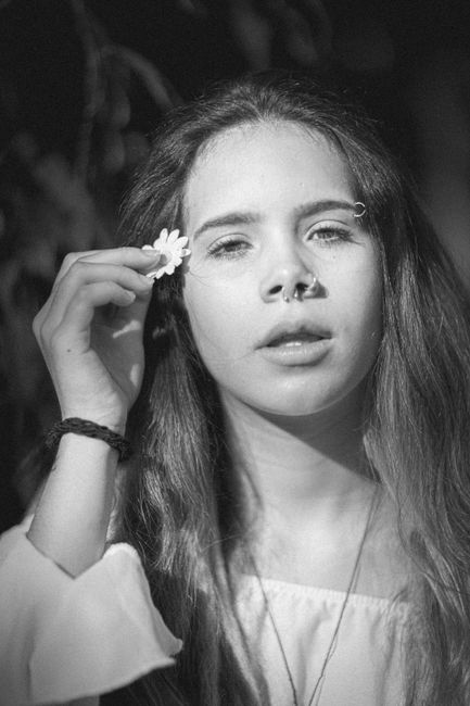 Black and white of calm female putting small flower behind ear and looking at camera while standing on blurred background