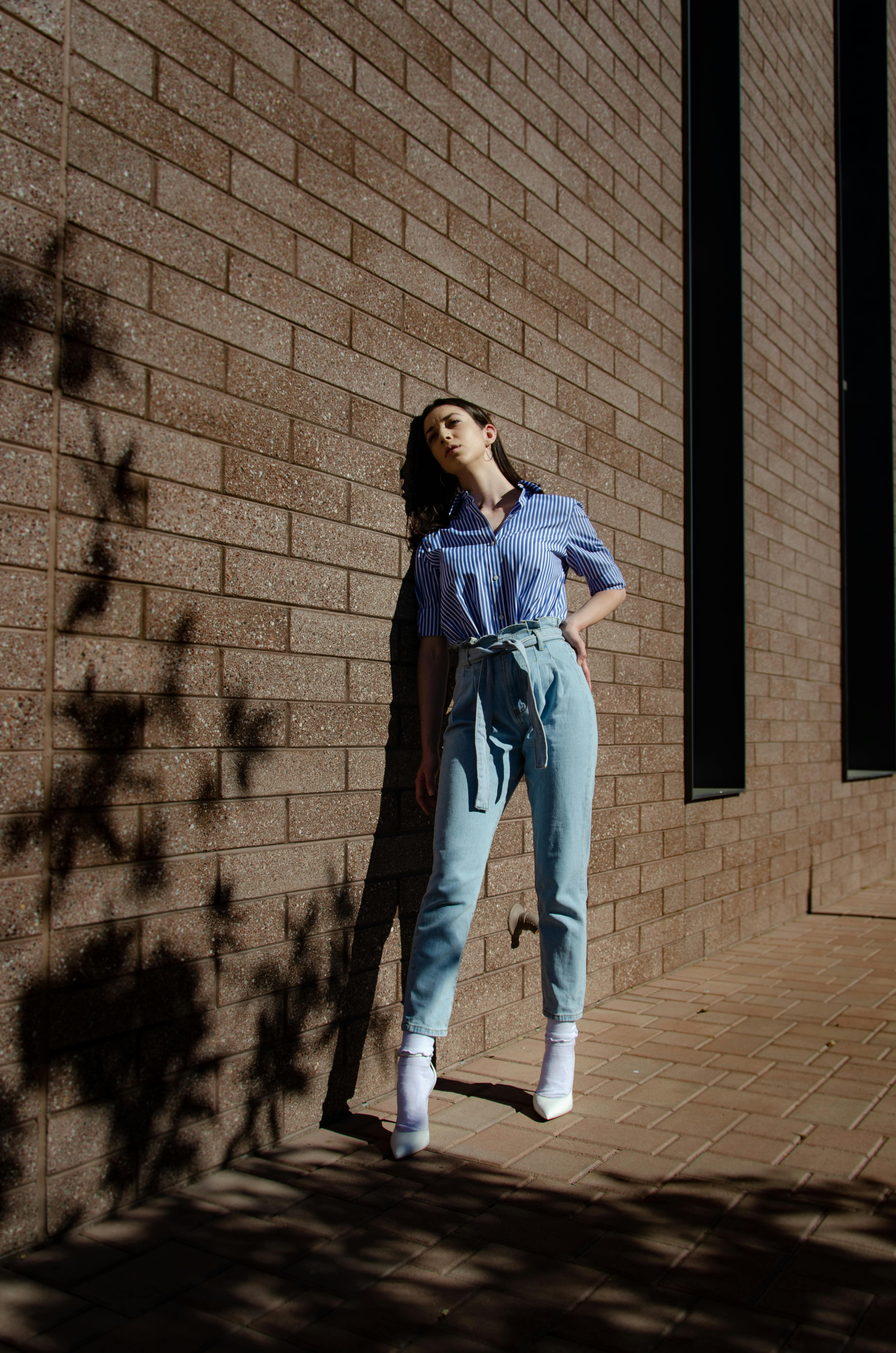 Full body of young woman in trendy outfit standing near building with brick wall on city street in sunny day