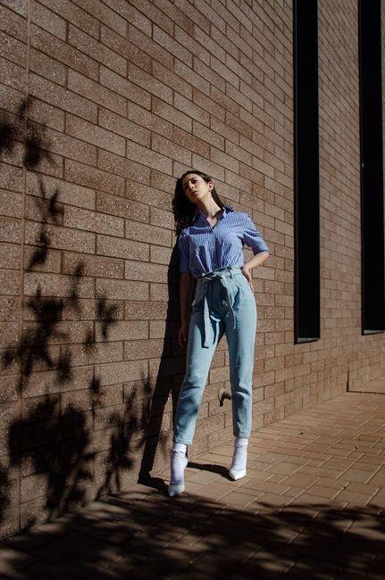 Full body of young woman in trendy outfit standing near building with brick wall on city street in sunny day