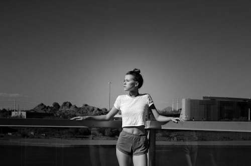 Black and white modern young lady in shorts and t shirt standing near glass fence and river with city on background in daytime and looking away