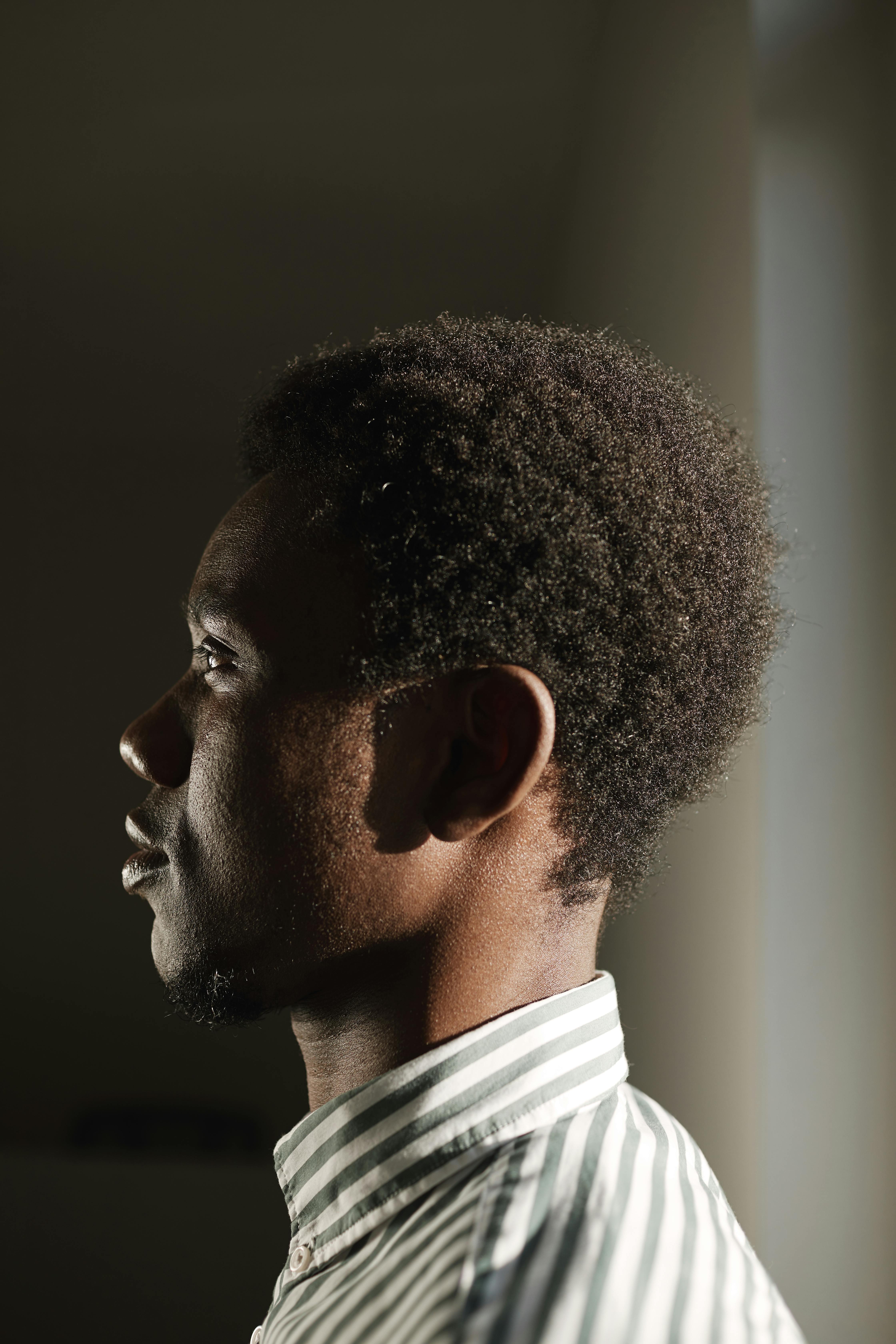Side profile of a young black man in striped shirt with soft lighting, thoughtful expression.