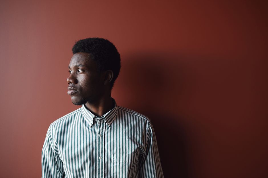 A young man in a striped shirt gazes thoughtfully against a warm red backdrop.