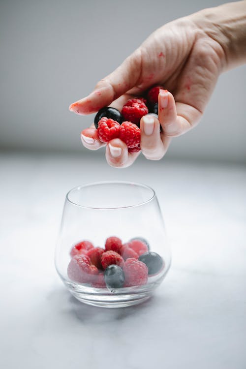 Crop anonymous female putting tasty raspberries and blueberries into glassware placed on white table