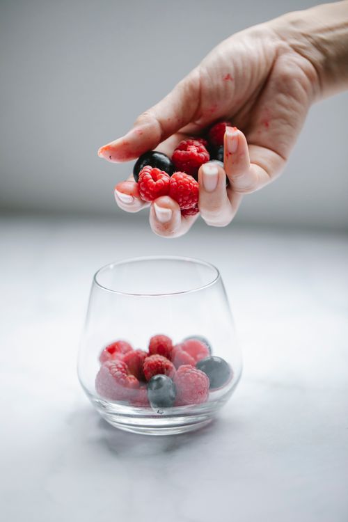 Free Crop anonymous female putting tasty raspberries and blueberries into glassware placed on white table Stock Photo
