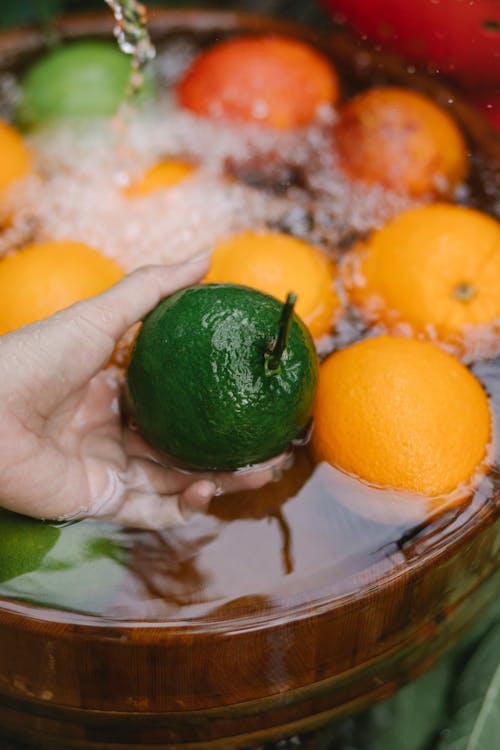 From above of crop anonymous person taking fresh green avocado from bowl with water and oranges