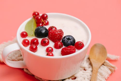 From above of delicious homemade yogurt in white ceramic mug decorated with assorted berries and served on pink table with wooden spoon