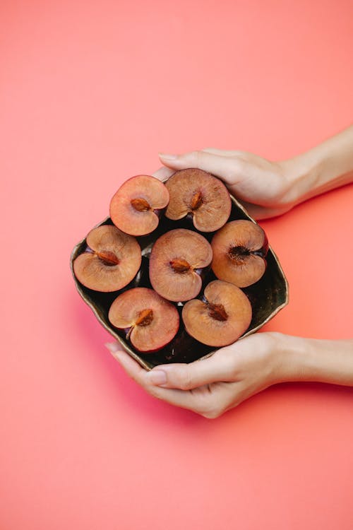 Top view of crop anonymous female serving bowl of fresh ripe halved plums on pink table